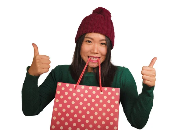 Young beautiful and happy Asian Korean woman in cool winter hat holding shopping bag excited buying Christmas present smiling in fashion beauty concept isolated — Stockfoto