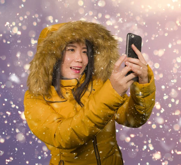Joven hermosa y feliz mujer asiática coreana en la capucha chaqueta de invierno tomando retrato selfie con el teléfono móvil al aire libre bajo la nieve sonriente alegre disfrutando de la Navidad — Foto de Stock