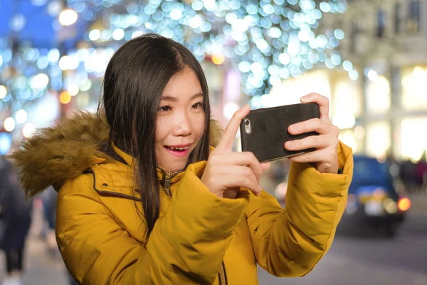 Mujer coreana asiática feliz en la capucha chaqueta de invierno tomando retrato selfie con el teléfono móvil al aire libre sonriendo alegre disfrutando de vacaciones de Navidad viaje y ciudad luces decoración — Foto de Stock