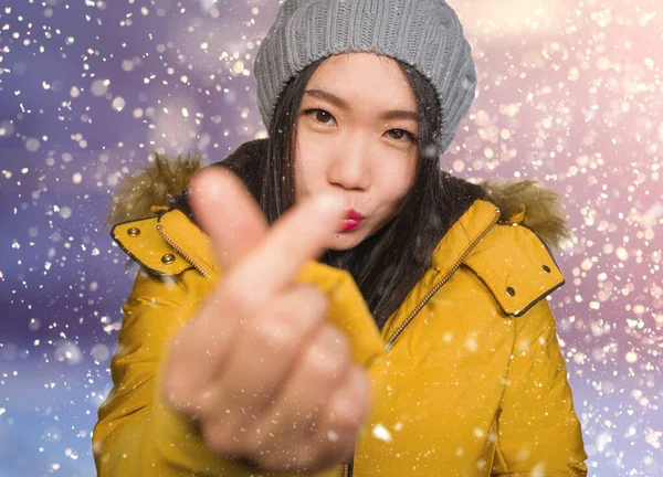 Young happy Asian Korean student woman in winter hat and warm hood jacket enjoying Christmas snowfall smiling cheerful and carefree  doing love finger sign — Stock Photo, Image