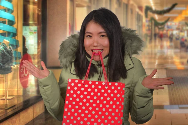 Joven feliz y hermosa mujer china asiática sosteniendo bolsa de compras roja comprar regalo de Navidad en el centro comercial de la ciudad sonriendo emocionado y alegre en abrigo de capucha de invierno caliente — Foto de Stock