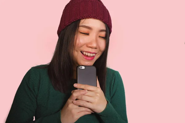 Joven hermosa y feliz asiática estudiante coreana utilizando aplicación de medios sociales en el teléfono móvil disfrutando emocionado y juguetón sonriente dulce usando sombrero de invierno aislado — Foto de Stock