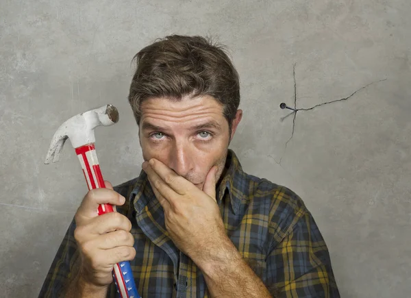 funny portrait of man holding hammer after driving a nail for hanging a frame but making a mess cracking the wall as a disaster DIY guy and messy domestic repair task