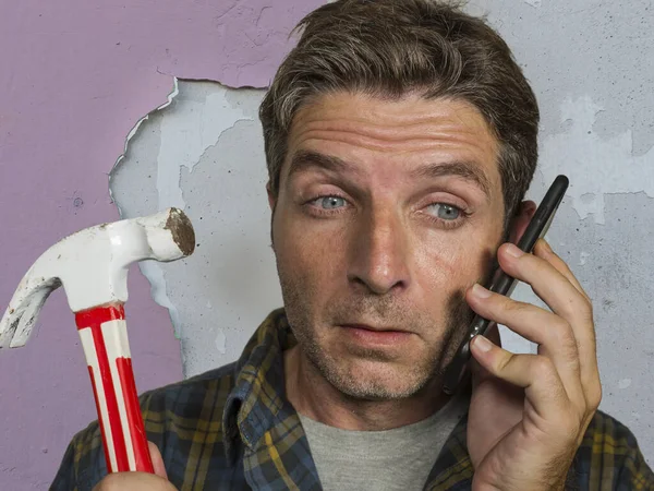 Retrato engraçado de homem confuso e frustrado chamando o seguro doméstico no telefone celular para a bagunça de reparo que ele fez com um martelo rachando a parede precisando de ajuda profissional — Fotografia de Stock