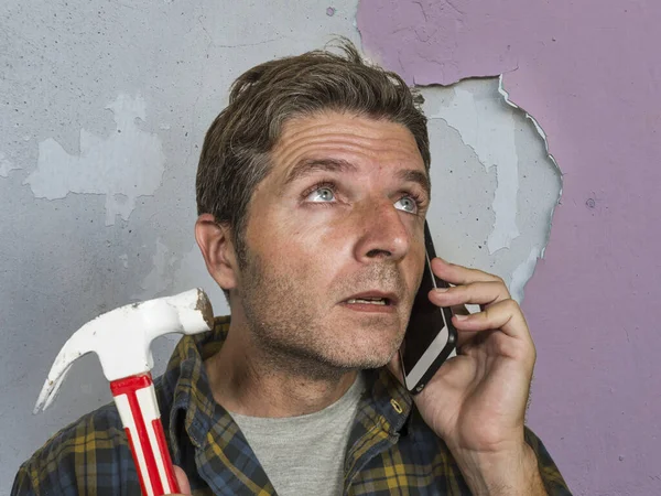 Lustiges Porträt eines chaotischen und frustrierten Mannes, der die Hausversicherung per Handy anruft, um das Chaos zu reparieren, das er mit einem Hammer angerichtet hat, der die Wand zertrümmert und professionelle Hilfe benötigt — Stockfoto