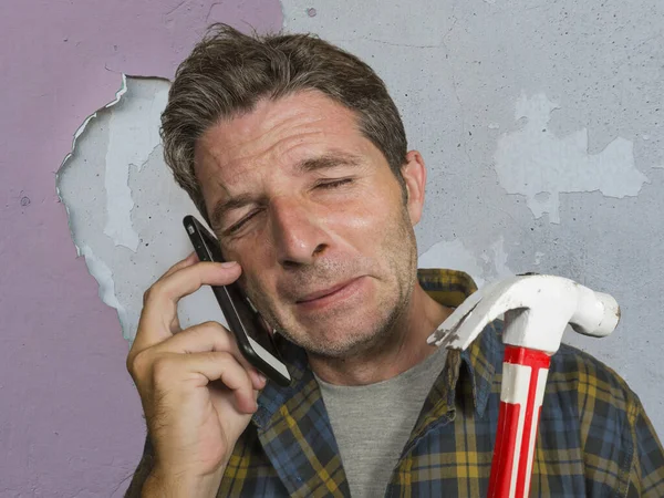 funny portrait of messy and frustrated man calling domestic insurance on mobile phone for repair mess he did with a hammer cracking the wall needing professional help