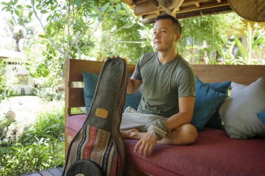 Genç, yakışıklı ve mutlu, hippi tarzlı, açık havada gitar çalan, sakin tropikal bahçede dinlenen, tatil inzivasının tadını çıkaran etnik kökenli bir adam.