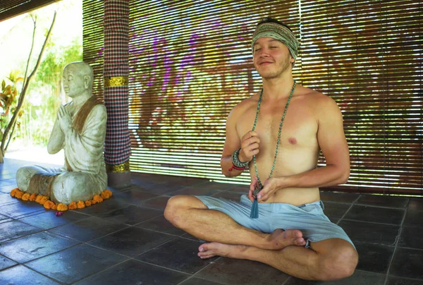 Jovem meditação hipster bonito sentado em posição de lótus - Attractive atlético e feliz homem praticando ioga relaxamento exercício vestindo faixa de cabelo e colar de contas mala — Fotografia de Stock