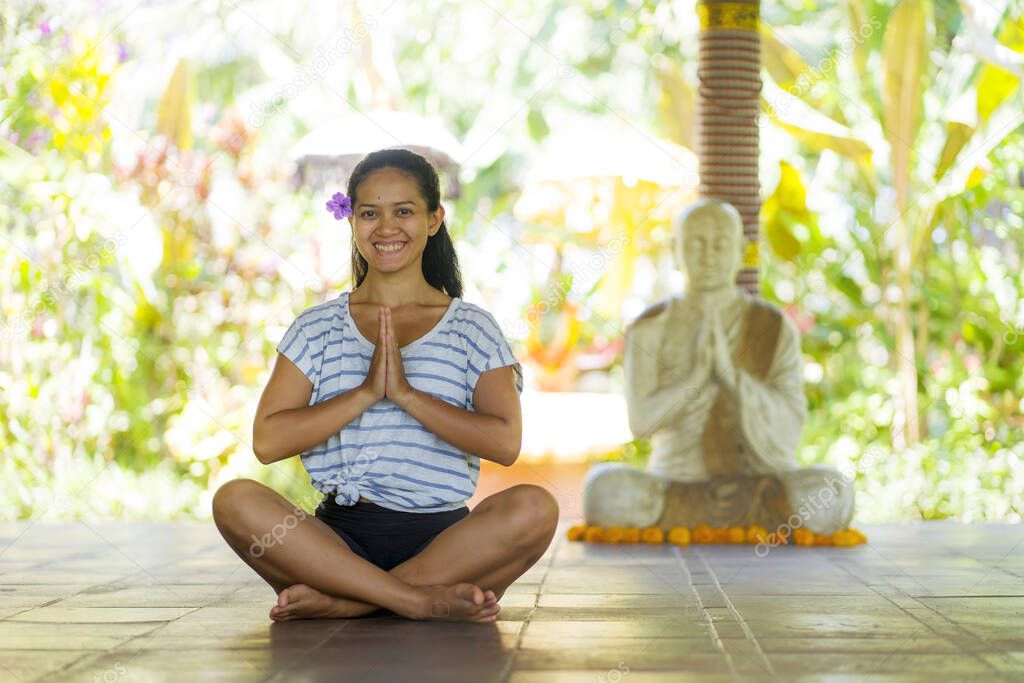 young beautiful and happy Asian Indonesian woman with Balinese style ear flower sitting on wood floor in lotus pose smiling at yoga studio relaxed and zen like in harmony