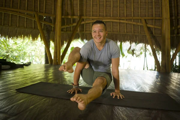 Junger attraktiver und glücklicher Mann, der Yoga-Übungen und Akro-Übungen macht, lächelt entspannt in Harmonie in einer wunderschönen asiatischen Bambushütte und genießt die Natur im körperlichen Gleichgewicht — Stockfoto