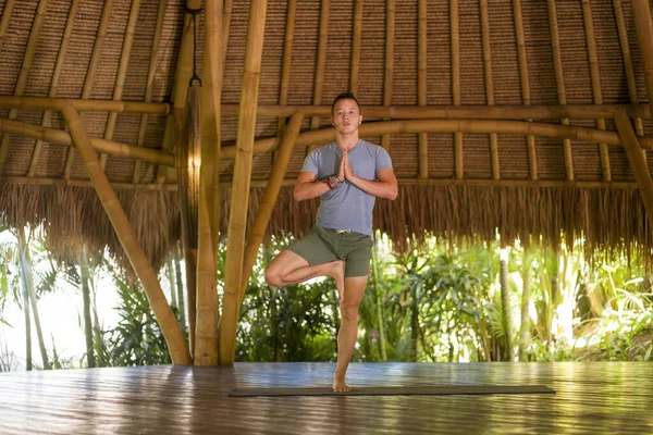 Junger attraktiver und glücklicher Mann bei Yoga-Übungen und Gleichgewichtsübungen entspannt in Harmonie in einer wunderschönen asiatischen Bambushütte, die Natur im körperlichen Gleichgewicht genießt — Stockfoto