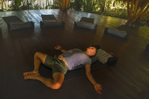 young attractive and happy man doing yoga drill lying on wood floor relaxed meditating in harmony at beautiful Asian bamboo hut enjoying nature in body and mind balance