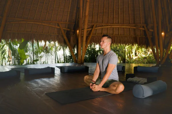 Junger attraktiver und glücklicher Mann macht Yoga sitzend in Lotusposition und meditiert entspannt in Harmonie in einer wunderschönen asiatischen Bambushütte und genießt die Natur im körperlichen und geistigen Gleichgewicht — Stockfoto