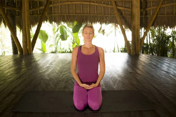 young happy and beautiful blond American woman doing yoga workout in Bali at exotic bamboo hut opened to the forest in fitness and healthy lifestyle enjoying retreat