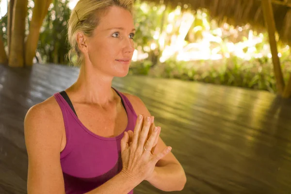 Beautiful blond American woman doing yoga workout in Bali at exotic bamboo hut opened to forest view sitting in namaste hands pose meditating enjoying retreat — Stock Photo, Image