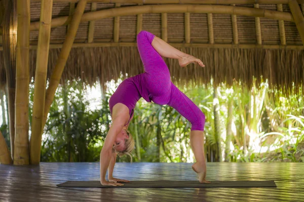 young happy and beautiful blond American woman doing yoga workout in Bali at exotic bamboo hut opened to the forest in fitness and healthy lifestyle enjoying retreat