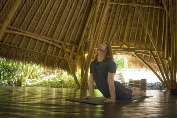 Joven atractivo y feliz hombre en estilo hipster yogui haciendo yoga taladro en la estera en hermosa cabaña de bambú asiático estiramiento cuerpo disfrutando de la naturaleza en equilibrio mental — Foto de Stock