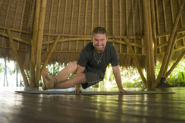 Jovem atraente e feliz homem no estilo hipster yogi fazendo ioga broca no tapete na bela cabana de bambu asiático alongamento corpo desfrutando da natureza em mente equilíbrio — Fotografia de Stock