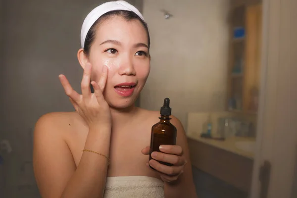 Естественный образ жизни портрет молодой красивой и счастливой азиатской корейской женщины, применяющей антивозрастную косметику сыворотки на лице в ванной комнате дома, улыбающейся свежей в уходе за кожей лица — стоковое фото