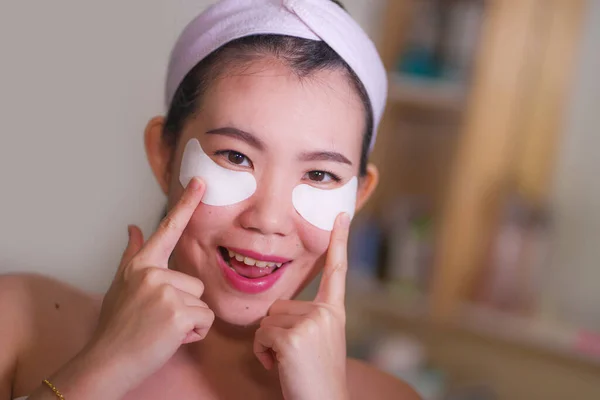 Молодая красивая и счастливая азиатская кореянка, улыбающаяся в зеркало ванной, нанося увлажняющие повязки под глазами в косметике и уходе за кожей лица — стоковое фото