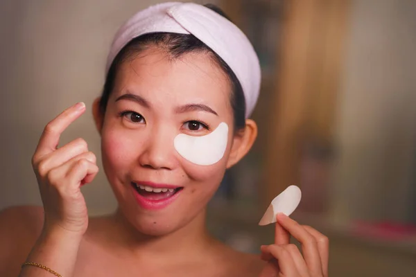Молодая красивая и счастливая азиатская китаянка улыбается в зеркало ванной, нанося увлажняющие повязки под глазами в косметике и уходе за кожей лица — стоковое фото