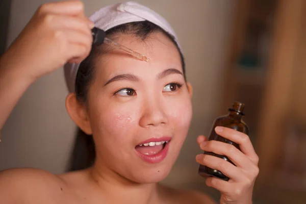 Estilo de vida natural retrato de la joven hermosa y feliz mujer coreana asiática aplicando suero anti envejecimiento cosmético en su cara en el baño en casa sonriendo fresco en el cuidado de la piel facial — Foto de Stock