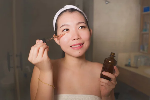 natural lifestyle portrait of young beautiful and happy Asian Korean woman applying anti aging serum cosmetic on her face at home bathroom smiling fresh in facial skin care