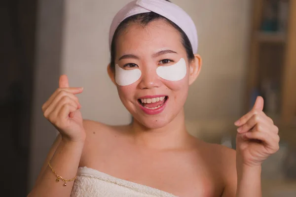 Молодая красивая и счастливая азиатка японка, улыбающаяся в зеркало ванной, наносила увлажняющие повязки под глазами в косметике и уходе за кожей лица — стоковое фото