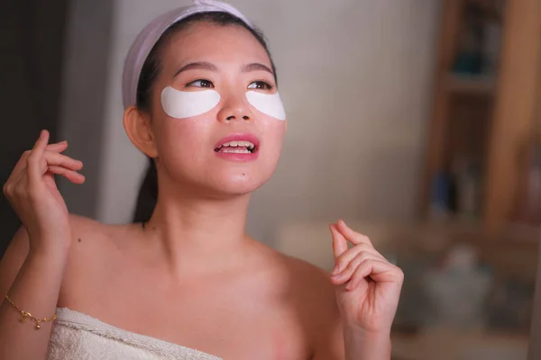 Молодая красивая и счастливая азиатская кореянка, улыбающаяся в зеркало ванной, нанося увлажняющие повязки под глазами в косметике и уходе за кожей лица — стоковое фото