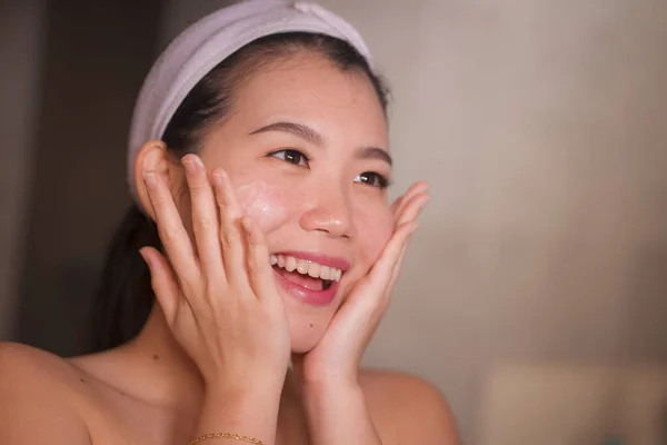 Stile di vita domestico ritratto di giovane donna cinese asiatica bella e felice che applica la crema cosmetica facciale sorridente allegra allo specchio del bagno nella cura della pelle di bellezza — Foto Stock