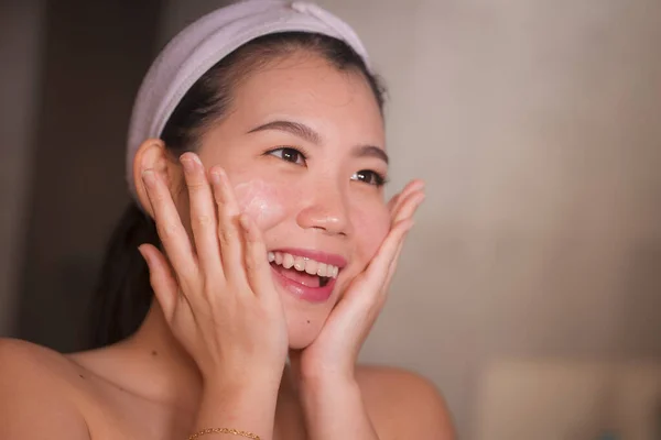 Retrato do estilo de vida doméstico da mulher chinesa asiática bonita e feliz nova que aplica o creme cosmético facial que sorri alegre ao espelho do banheiro no cuidado da pele da beleza — Fotografia de Stock