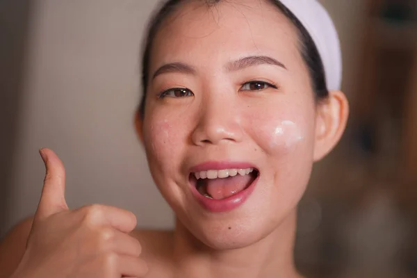 Estilo de vida doméstico retrato de joven hermosa y feliz mujer china asiática aplicando crema cosmética facial sonriente alegre al espejo de baño en el cuidado de la piel belleza — Foto de Stock