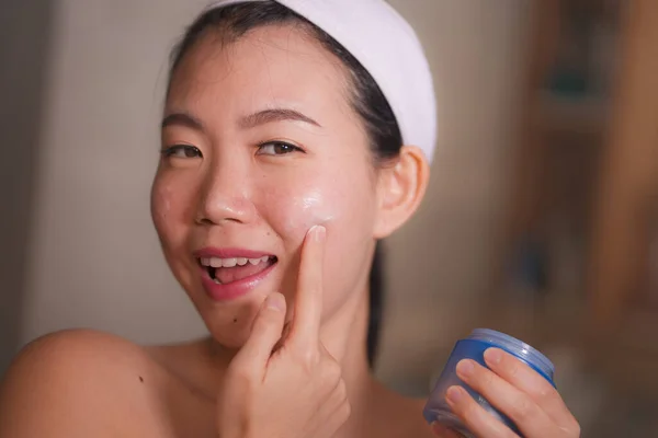 Retrato do estilo de vida doméstico da mulher chinesa asiática bonita e feliz nova que aplica o creme cosmético facial que sorri alegre ao espelho do banheiro no cuidado da pele da beleza — Fotografia de Stock