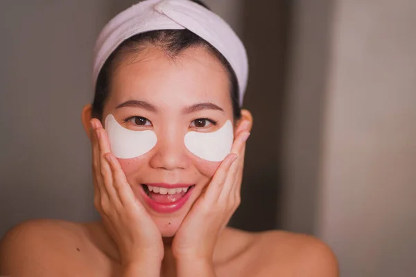 Wanita muda Asia yang cantik dan bahagia tersenyum pada cermin kamar mandi menerapkan tambalan mata yang terhidrasi di bawah mata dalam kosmetik kecantikan dan perawatan kulit wajah — Stok Foto