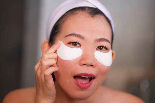 Wanita muda Cina cantik dan bahagia tersenyum ke kamar mandi cermin menerapkan tambalan mata terhidrasi di bawah mata dalam kosmetik kecantikan dan perawatan kulit wajah — Stok Foto