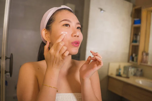 Retrato de estilo de vida doméstico de jovem bonita e feliz mulher asiática coreana aplicando creme cosmético facial sorrindo brincalhão ao espelho do banheiro em cuidados com a pele de beleza — Fotografia de Stock
