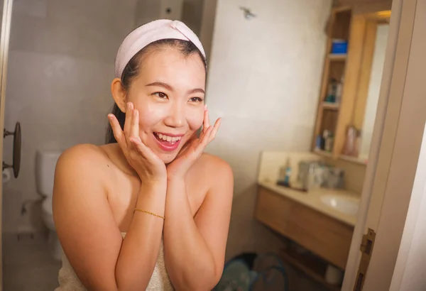 Retrato do estilo de vida doméstico da mulher chinesa asiática bonita e feliz nova que aplica o creme cosmético facial que sorri alegre ao espelho do banheiro no cuidado da pele da beleza — Fotografia de Stock