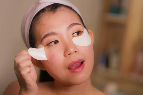 Jovem bela e feliz mulher chinesa asiática sorrindo para espelho do banheiro aplicando manchas olho hidratante sob os olhos em cosméticos de beleza e cuidados com a pele facial — Fotografia de Stock