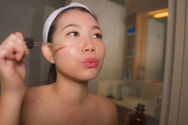 Estilo de vida natural retrato de jovem bonita e feliz mulher asiática coreana aplicando anti envelhecimento soro cosmético em seu rosto em casa casa de banho sorrindo fresco em cuidados com a pele facial — Fotografia de Stock