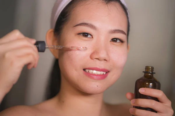 年轻、美丽、快乐的亚裔女性在家里的浴室里用抗衰老血清化妆品在脸上挂上自然的生活方式画像，脸上带着新鲜的面部肌肤护理的微笑 — 图库照片