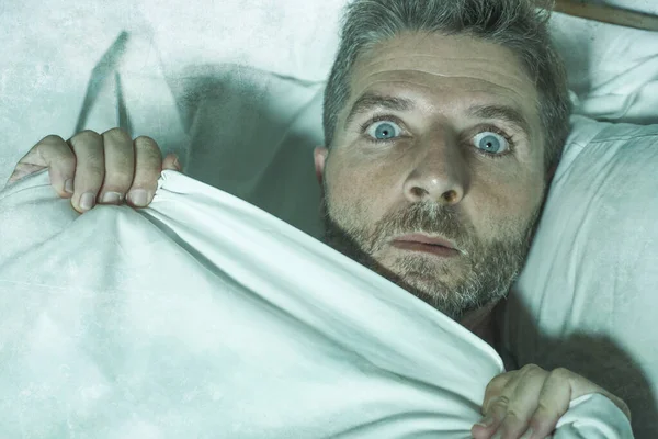 Напряженный и напуганный мужчина один в постели просыпается ночью в страхе после кошмара, чувствуя паранойю держа одеяло в смешной выражение панического лица — стоковое фото