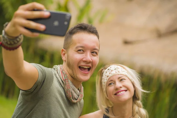 Joven feliz y hermosa pareja hipster tomando foto selfie con teléfono móvil al aire libre disfrutando de vacaciones en el fondo de la naturaleza tropical sonriendo y explorando — Foto de Stock