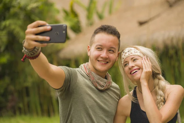 Молодая счастливая и красивая пара хипстеров делает селфи-фото с мобильного телефона на открытом воздухе наслаждаясь отдыхом на тропическом фоне природы улыбаясь и исследуя — стоковое фото