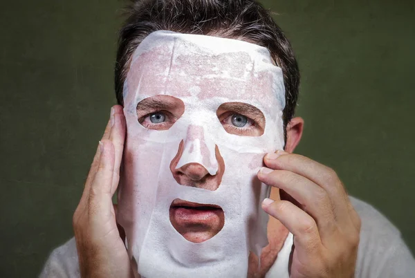 lifestyle isolated background portrait of young weird and funny man at home trying using paper facial mask cleansing applying anti aging beauty treatment