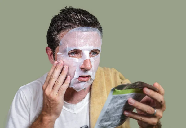 Νεαρός περίεργος και αστείος άνθρωπος στο σπίτι προσπαθεί να χρησιμοποιήσει χαρτί καθαρισμού μάσκα προσώπου εφαρμογή αντι γήρανση θεραπεία προσώπου ανάγνωση οδηγίες προϊόν ομορφιάς — Φωτογραφία Αρχείου