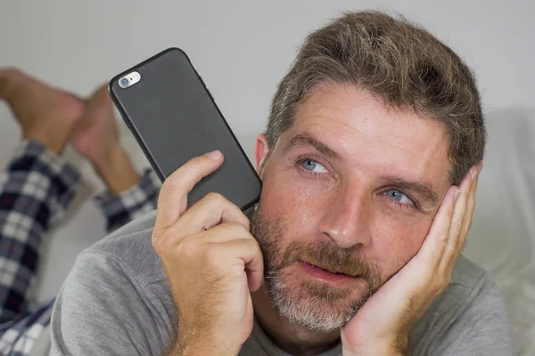 Retrato do estilo de vida doméstico de homem jovem pensativo e atraente deitado na cama em casa pensando e sonhando acordado relaxado usando rede de telefone móvel vestindo pijama — Fotografia de Stock