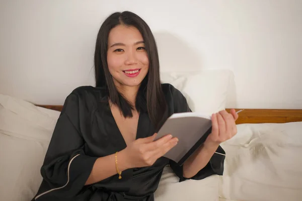 Молодая красивая и милая азиатская кореянка в постели читает книгу роман чувствуя себя счастливым и расслабленным или учиться дома в ночнушке выглядит уютно и уютно — стоковое фото