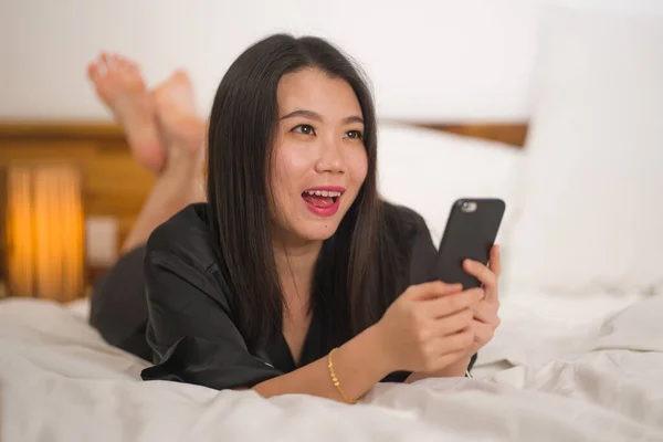 身穿可爱睡衣的年轻漂亮甜美的亚裔韩国女人在床上玩手机，享受社交媒体或网上约会应用的快乐和放松 — 图库照片