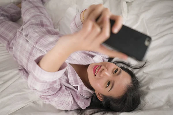 Joven hermosa y dulce mujer coreana asiática en pijama lindo juguetón en la cama usando el teléfono móvil disfrutando de las redes sociales o en línea citas aplicación sonriendo feliz y relajado — Foto de Stock