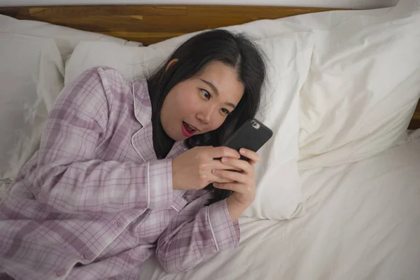 身穿可爱睡衣的年轻漂亮甜美的亚裔韩国女人在床上玩手机，享受社交媒体或网上约会应用程序的快乐和放松 — 图库照片